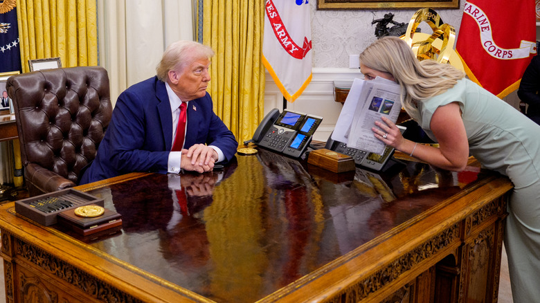 Karoline Leavitt se penche sur le bureau de Donald Trump et cachant sa bouche derrière un journal tout en lui parlant
