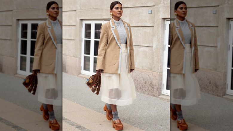 Zufi Alexander porte une jupe transparente blanche, des chaussettes grises, des mules marron, un haut gris, une veste marron et un sac en daim marron avec breloques lors de la Fashion Week de Copenhague