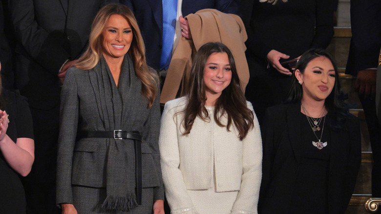 Melania Trump debout et souriante dans un manteau gris avec un foulard