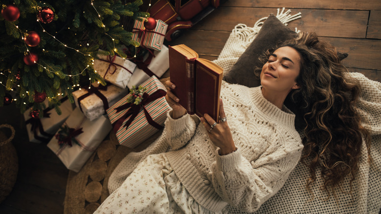 Jeune femme lisant un livre, allongée près de l'arbre de Noël dans une pièce chaleureuse et décorée