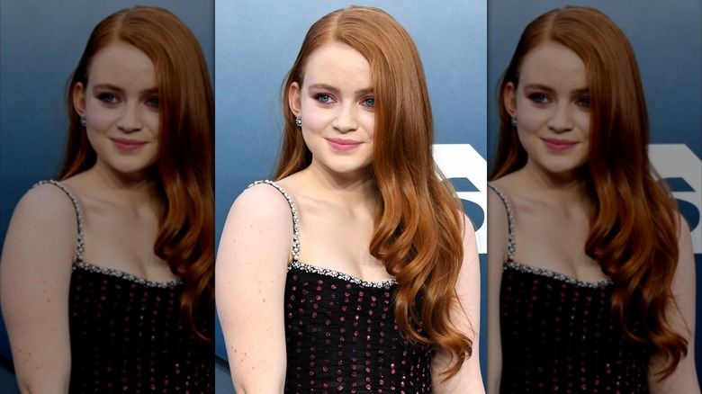 Sadie Sink souriante avec une raie sur le côté avec des boucles lâches à la fin