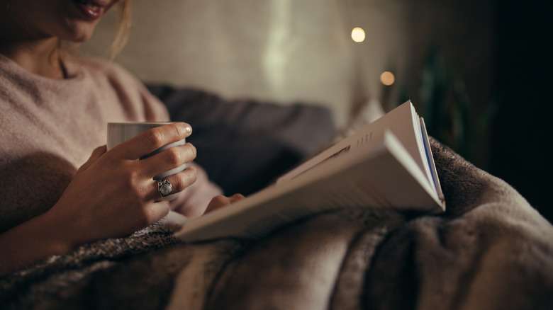 Une femme assise sur un canapé avec une tasse de thé et lisant un livre