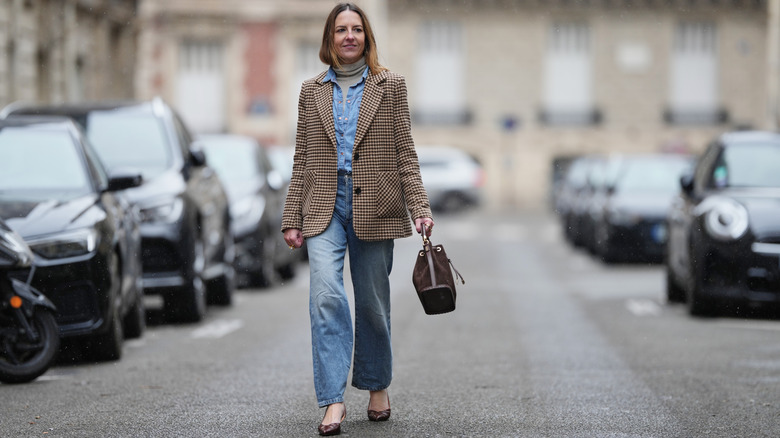 Alba Garavito Torre porte de longs cheveux châtain clair détachés avec une raie centrale, un maquillage naturel avec des sourcils définis et un rouge à lèvres doux, un pull à col roulé beige, une chemise en jean bleu clair superposée, une veste blazer à motif pied-de-poule beige et marron.