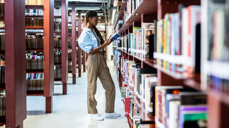 Plan long d'une femme avec une coiffure tressée et un pantalon kaki parcourant des livres dans une bibliothèque