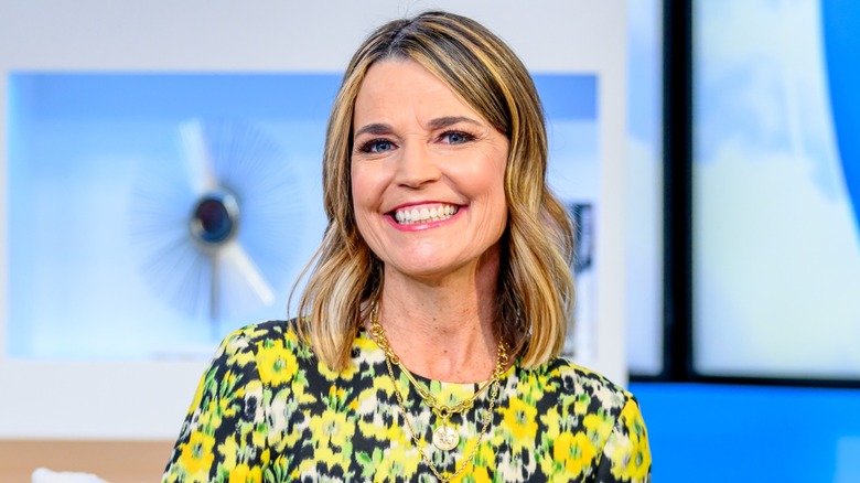 Savannah Guthrie souriante dans un studio de télévision portant un haut à imprimé floral jaune, blanc et noir