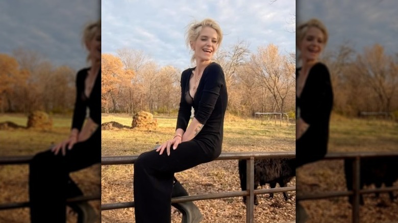 Kelly Osbourne sourit vêtue d'une robe noire alors qu'elle est assise sur une clôture