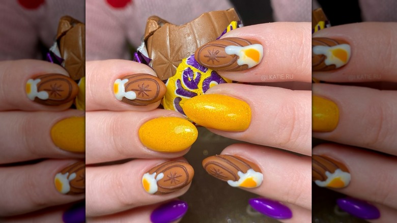 Gros plan de deux mains tenant des œufs à la crème Cadbury partiellement mangés tout en ayant un design d'ongle inspiré du bonbon