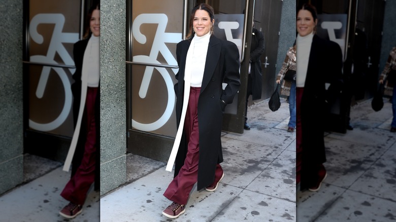 Neve Campbell marchant dans la rue avec des baskets assorties à son pantalon, un haut blanc et un manteau noir