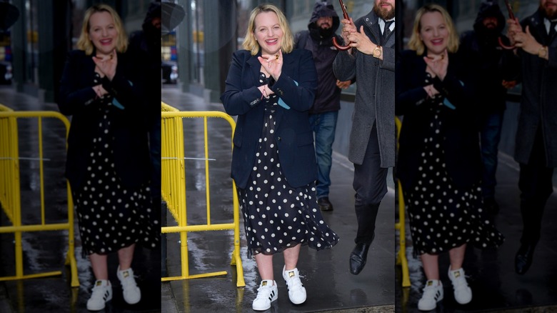 Elisabeth Moss marchant sous la pluie dans une robe à pois noir et blanc avec un blazer bleu marine et des baskets