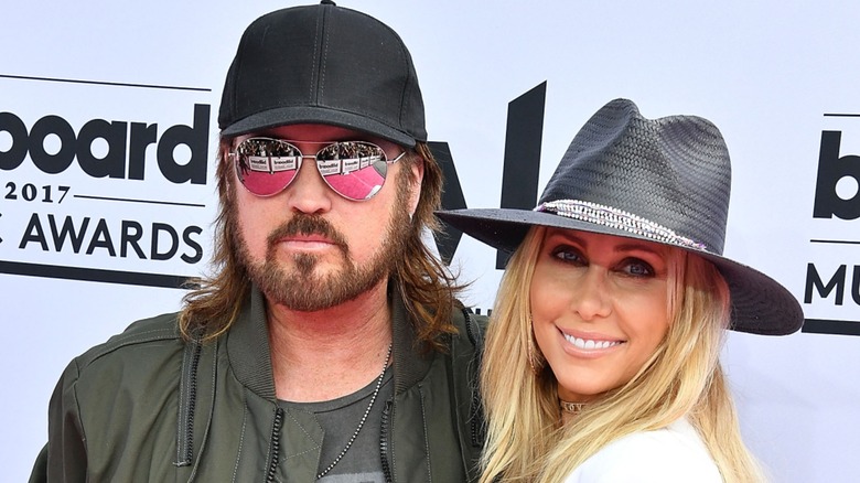 Billy Ray Cyrus avec un chapeau et des lunettes de soleil aux côtés de Tish Cyrus dans un fedora sur le tapis rouge