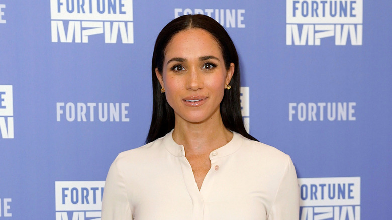 Meghan Markle sur le tapis rouge avec les cheveux raides et vêtue d'une robe chemise blanche