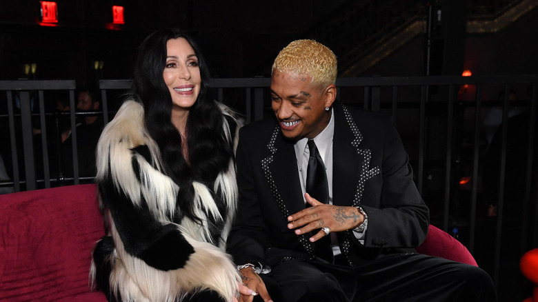 Cher sourit portant un manteau de fourrure noir et blanc à côté d'Alexander Edwards dans un blazer orné