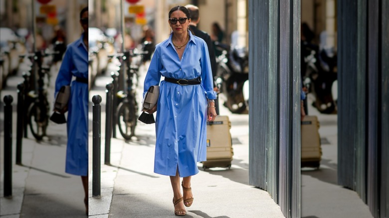 Une femme vêtue d'une robe chemise bleue marchant dehors avec des lunettes de soleil et une ceinture noire