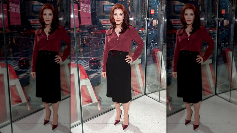 Priscilla Presley portant un bouton rouge et une jupe noire tout en posant devant une fenêtre de la ville
