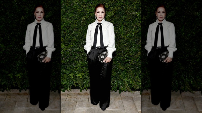 Priscilla Presley posant devant un mur de haie tout en portant une chemise blanche et un pantalon en satin