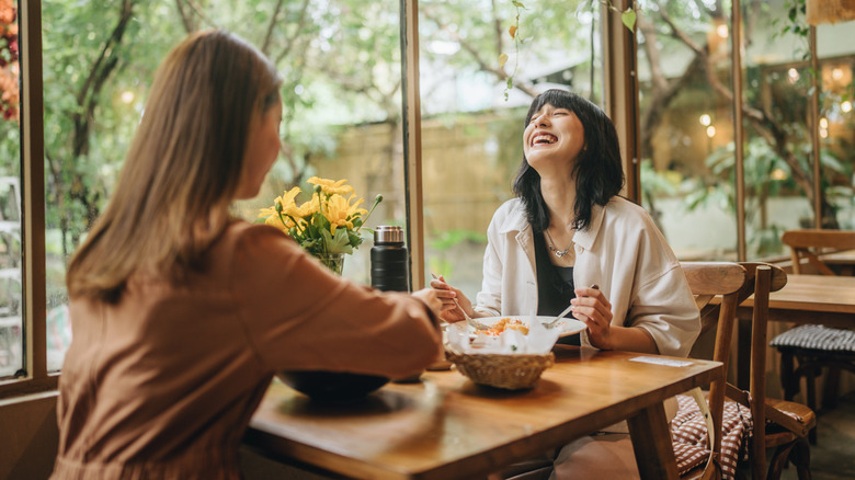 Deux femmes prennent un repas ensemble et rient en s'asseyant près d'une grande fenêtre