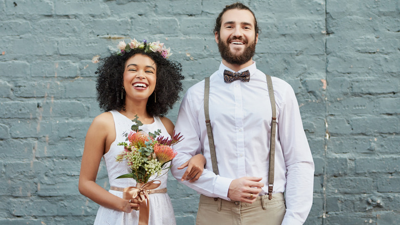 Les mariés posent contre des briques bleues, la mariée portant une couronne de fleurs tandis que le marié porte des bretelles et un nœud papillon