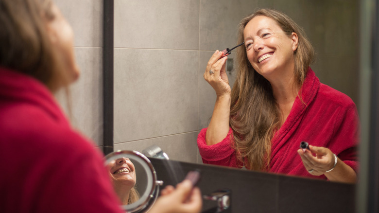 Le reflet heureux d'une femme âgée vu dans un miroir alors qu'elle applique du mascara tout en portant une robe rouge