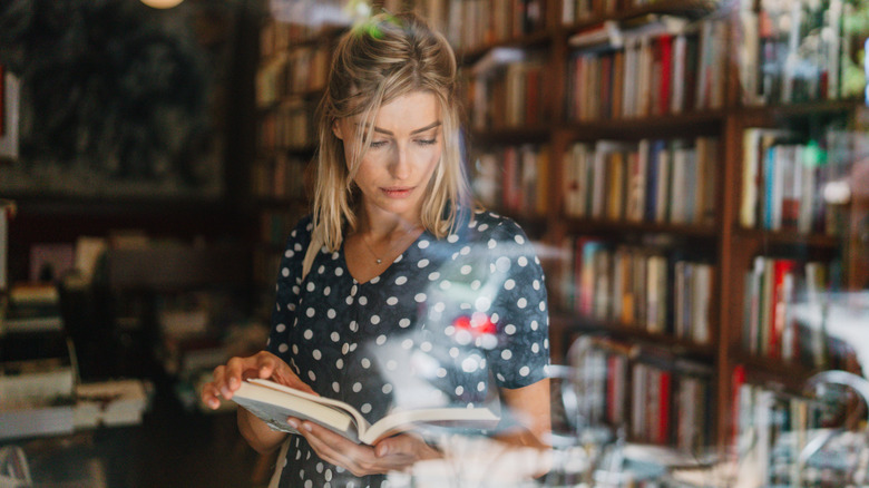 Une femme vêtue d'une robe bleue à pois lisant un livre dans une librairie à travers une fenêtre