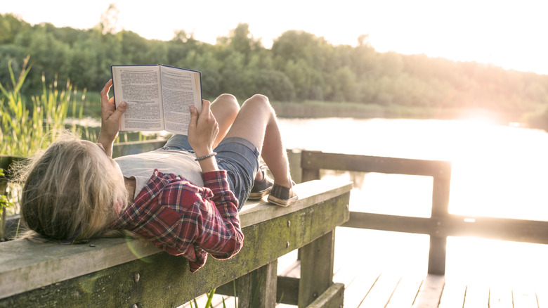 jeune fille lisant un livre en étant allongée sur le dos sur une poutre en bois sur un quai en bois au bord du lac