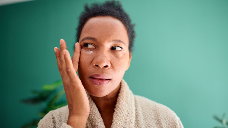 Une femme en peignoir devant un fond vert appliquant une crème pour les yeux et regardant sur le côté
