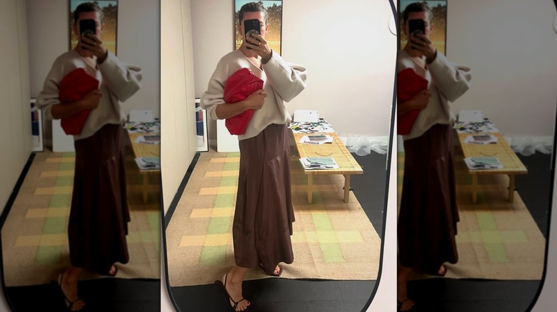 Une femme prenant un selfie miroir d'elle-même dans une jupe marron, un pull crème et des sandales noires tout en tenant une pochette rouge