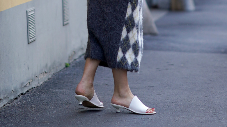 Vue latérale de petits talons blancs comportant une grande bride sur le pied, portés avec une jupe grise et à carreaux