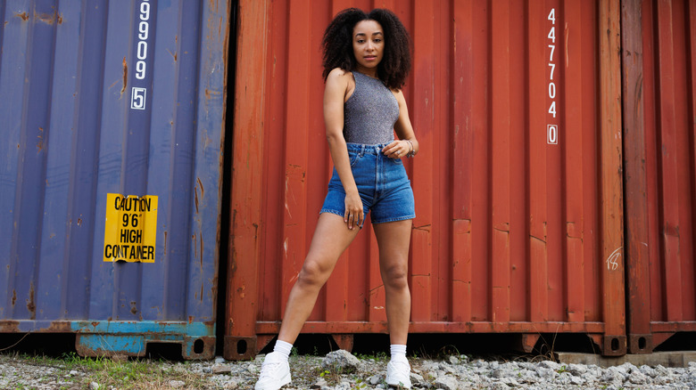 Jeune femme aux cheveux bouclés posant devant des conteneurs de fret tout en portant un short en jean taille haute et ajusté