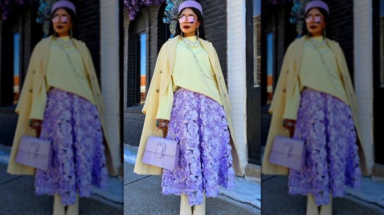 Femme en pull jaune beurre, chaussures et manteau avec jupe lavande, lunettes de soleil, chapeau et sac à main.