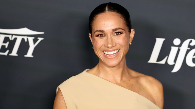 Meghan Markle souriante dans une robe crème à une épaule avec les cheveux lissés en arrière