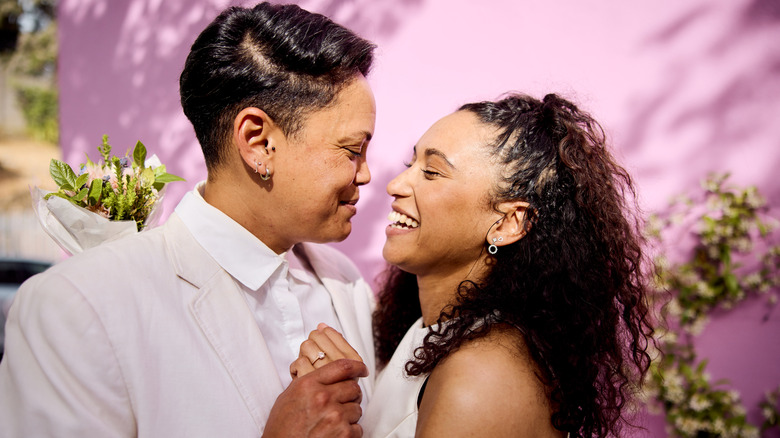 Couple queer souriant alors qu'ils s'embrassent en tenue de mariage blanche devant un bâtiment rose