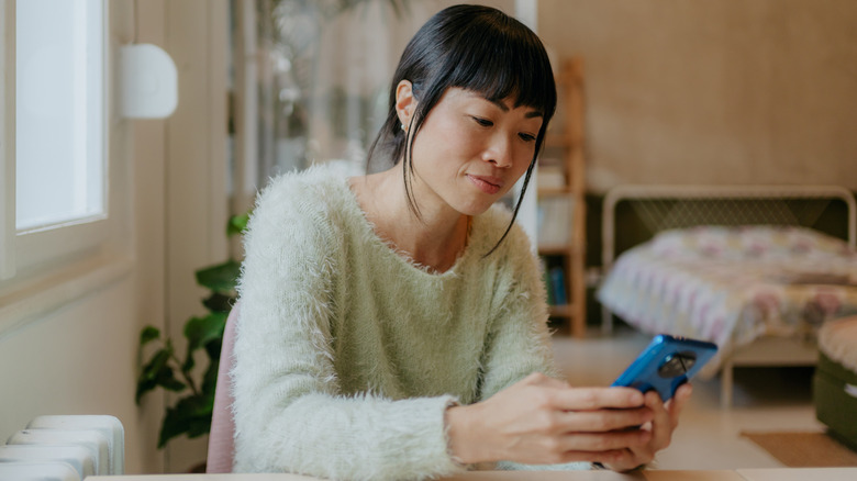 Femme dans un pull pelucheux se concentrant sur son téléphone intelligent bleu à la maison