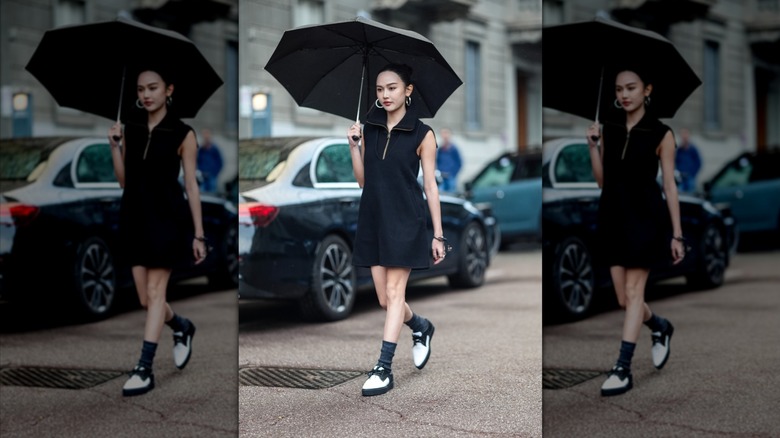 Une femme marchant dans la rue avec un parapluie, une mini-robe noire, un bandeau et des chaussures derby blanches