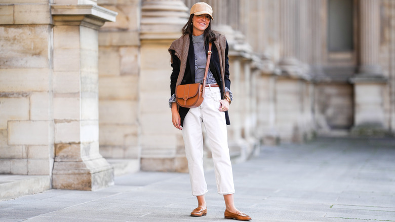 Une femme en chaussures plates et un pantalon crème avec une veste et un pull sur les épaules