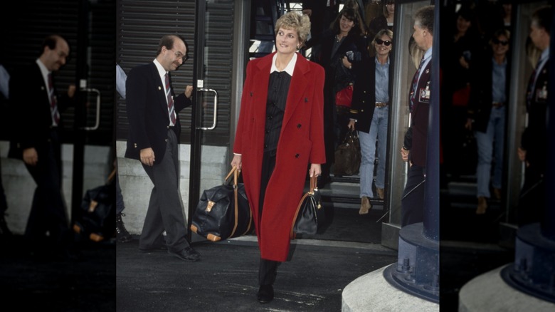 La princesse Diana porte un long manteau rouge alors qu'elle transporte des sacs dans un aéroport