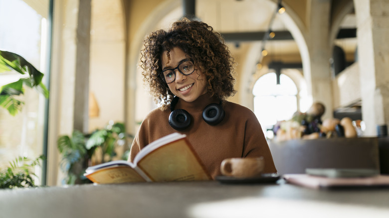 Une femme assise à une table avec une tasse de café tout en lisant et en réservant et portant des écouteurs autour du cou