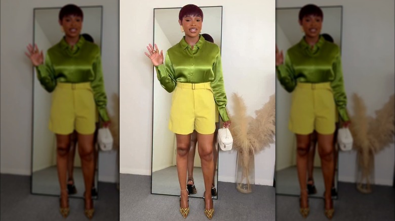 Une femme debout près d'un miroir dans une chemise en satin vert olive avec un short taille haute jaune beurre