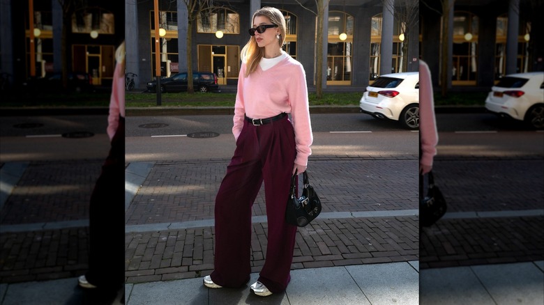 Une femme portant un pull rose pétale avec un pantalon bordeaux tout en posant dans la rue