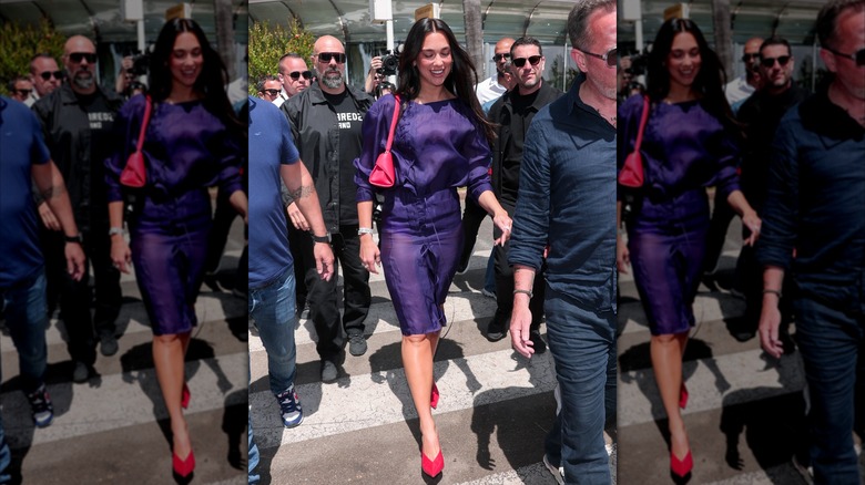 Dua Lipa marchant dans une foule vêtue d'une robe violette avec un sac et des chaussures rouges