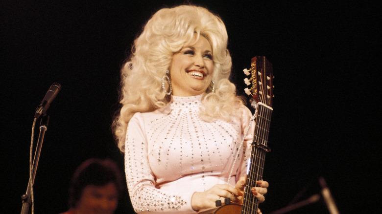 La jeune Dolly Parton rit sur scène tout en portant un costume blanc et une grande perruque bouclée
