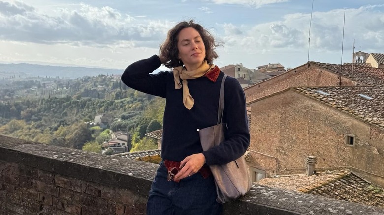Emma Ferrer posant les yeux fermés alors qu'elle s'appuie contre une clôture en brique avec un pittoresque village italien en arrière-plan