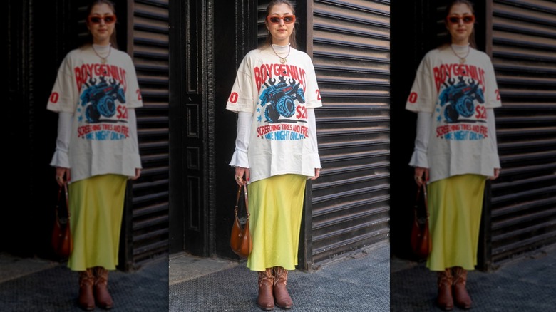 Une femme debout dans la rue vêtue d'une jupe midi jaune, d'un t-shirt surdimensionné et de bottes de cowboy