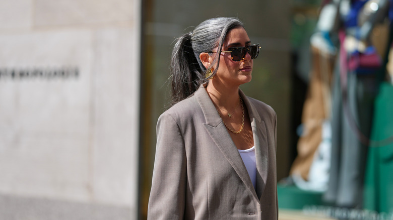 Une femme avec une queue de cheval grise et une veste aux tons champignon avec des lunettes de soleil à l'extérieur