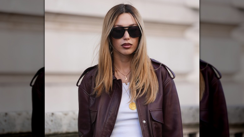 Femme aux cheveux blonds et aux racines plus foncées avec une partie centrale, des lunettes de soleil carrées noires surdimensionnées avec des verres foncés, un blouson aviateur en cuir bordeaux, un débardeur côtelé blanc