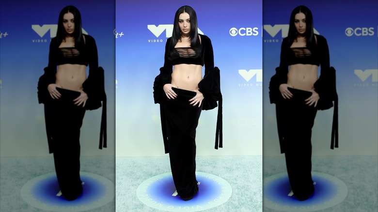 Charli XCX accrochant ses pouces dans le haut de sa jupe taille basse tout en portant un ensemble deux pièces
