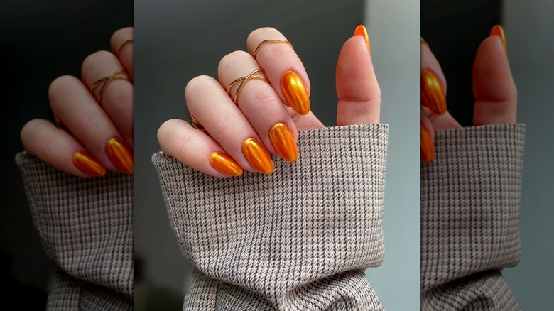 Main généralement fermée montrant des ongles orange avec une finition chromée ainsi qu'une pochette avec un motif en tweed
