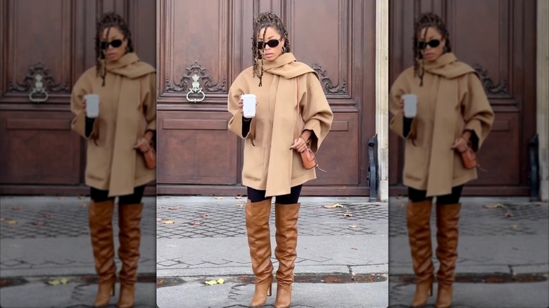 Une femme vêtue d'une veste camel avec des bottes souples camel tenant un café