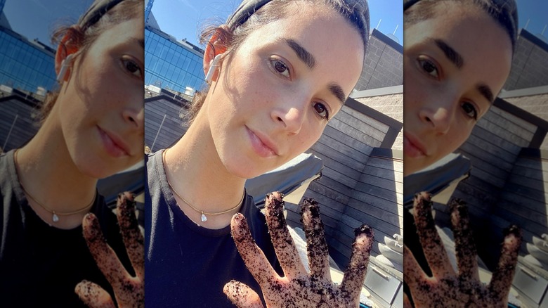 Aly Raisman avec ses cheveux attachés en arrière et un écouteur sans fil dans l'oreille levant une main avec de la terre dessus