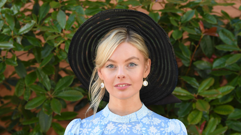 Nell Hudson posant devant de la verdure portant un chapeau noir et une robe bleue