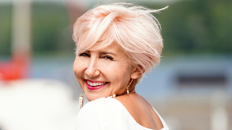 Une femme plus âgée avec une coupe bixie rose clair sourit à la caméra